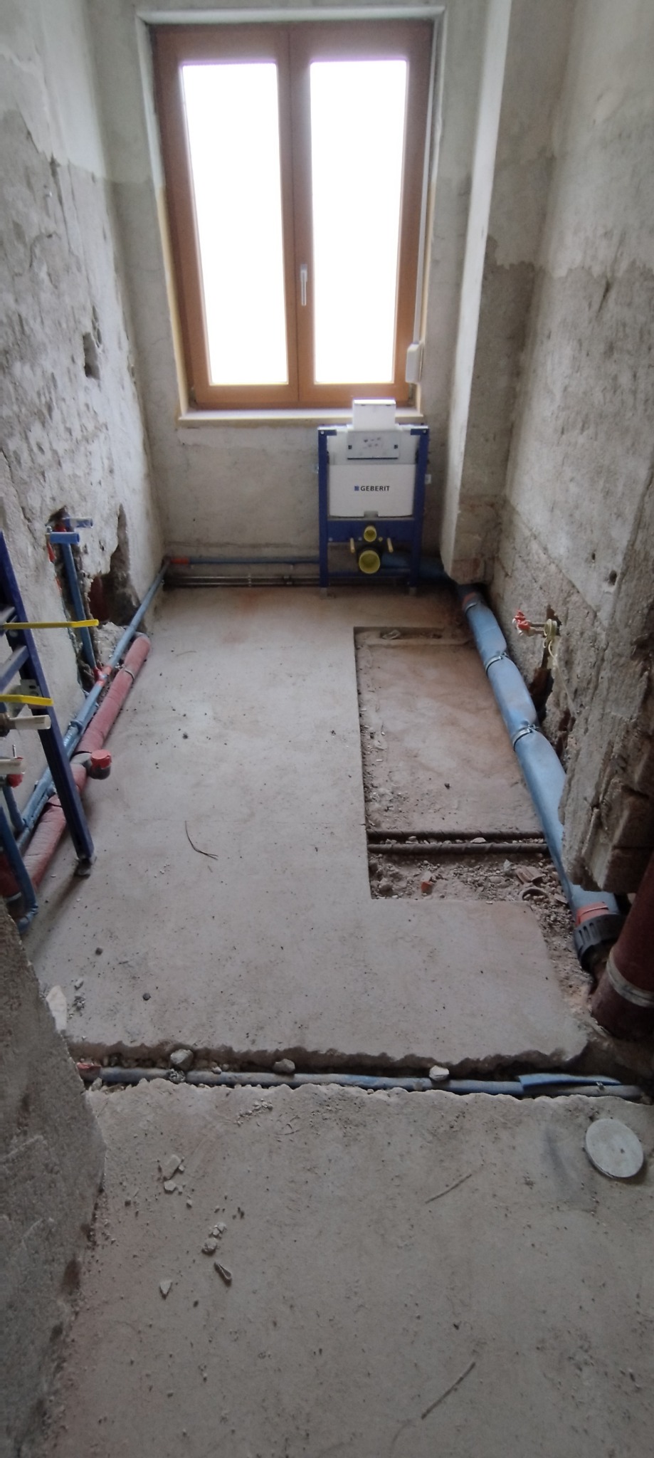 Surowy stan łazienki w trakcie remontu: widoczne instalacje wodne i kanalizacyjne, stelaż podtynkowy Geberit, ściany w stanie deweloperskim z widocznymi fragmentami cegieł, okno w tle.