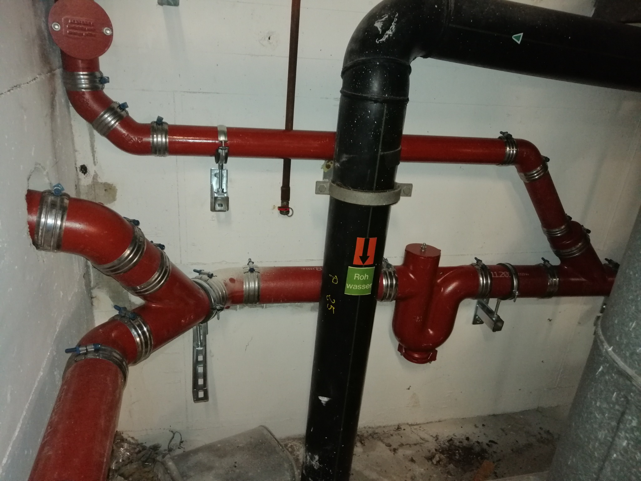 Czerwone i czarne rury instalacji wodnej z metalowymi obejmami zamocowane do betonowej ściany, widoczny fragment instalacji z zaworem i napisem 'Roh wasser'.