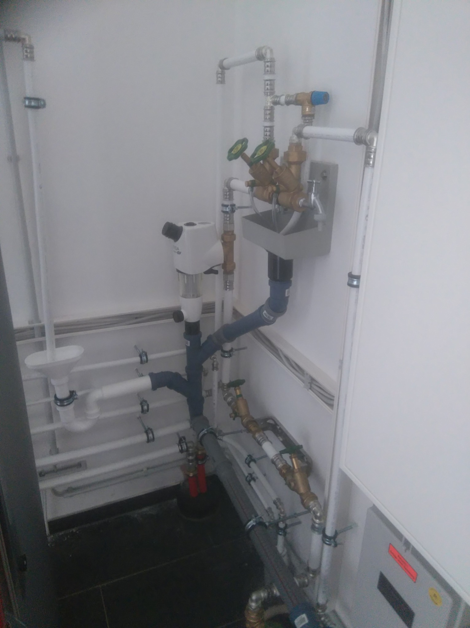 Kompleksowa instalacja hydrauliczna z białymi rurami PCV, zaworami z zielonymi pokrętłami, filtrem wody z przezroczystym zbiornikiem i odpływem lejkowym, zamontowana w pomieszczeniu z białą ścianą...