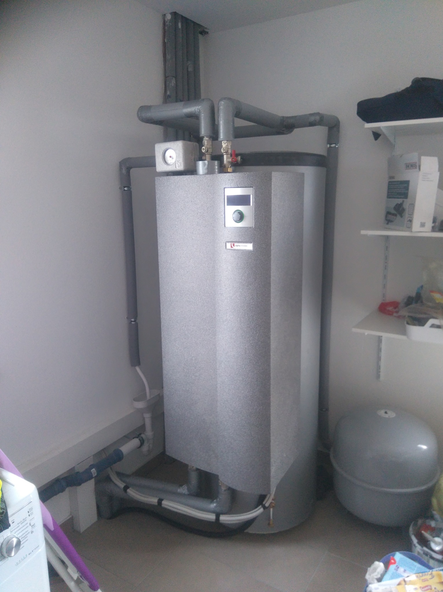 Szary, pionowy zasobnik ciepłej wody z podłączonymi rurami, widoczny manometr i panel sterowania, obok szare naczynie wzbiorcze, w tle półki z różnymi przedmiotami.