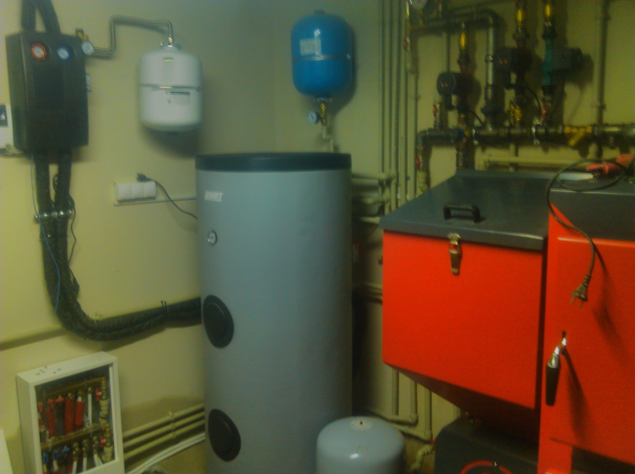 Kotłownia z szarym zasobnikiem ciepłej wody, czerwonym kotłem na ekogroszek, białym i niebieskim naczyniem przeponowym oraz rozdzielaczem instalacji grzewczej w białej obudowie.