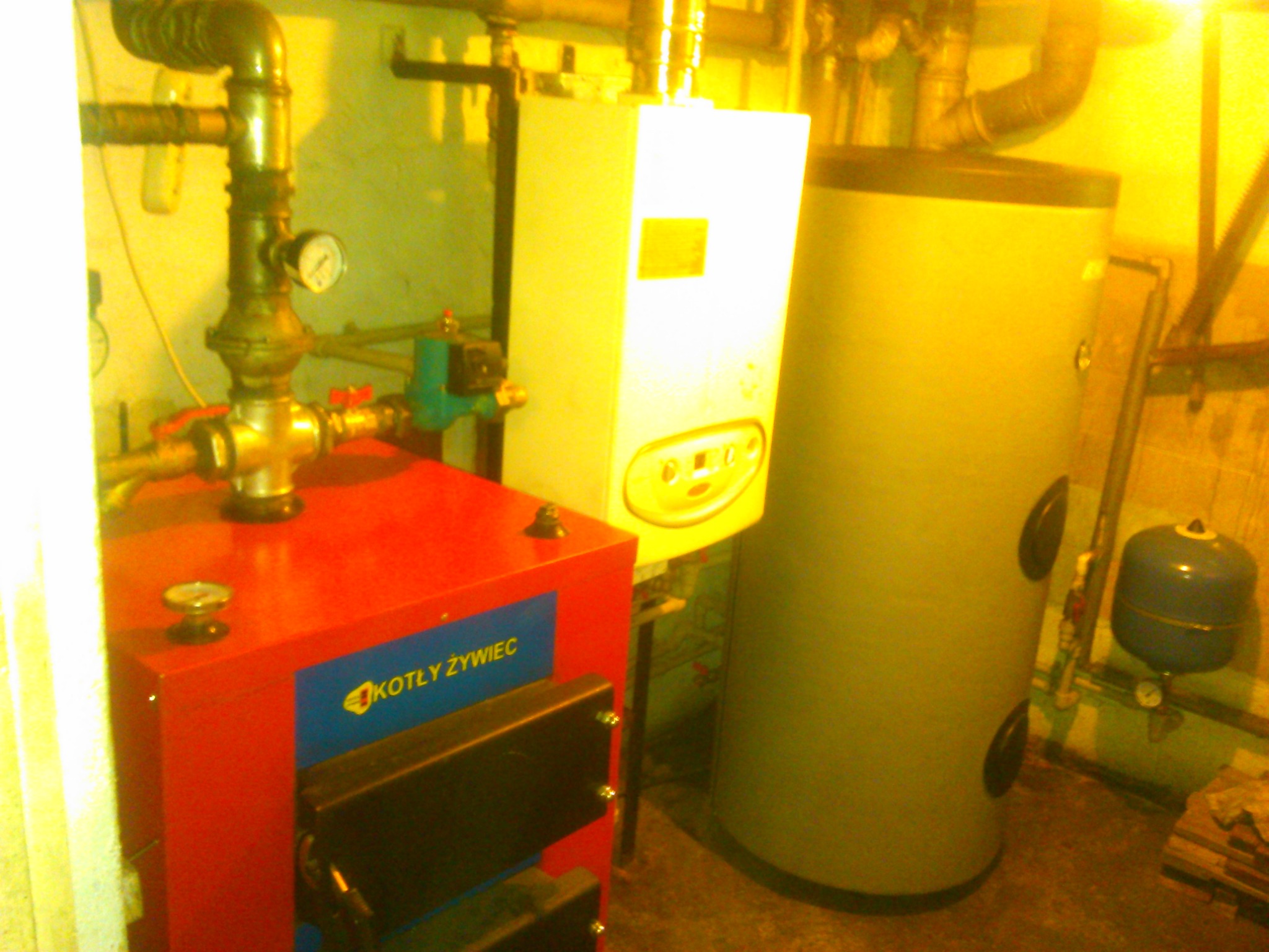 Czerwono-niebieski kocioł marki Kotły Żywiec, obok biały piec gazowy i szary zasobnik ciepłej wody w pomieszczeniu kotłowni, widoczne rury i zawory.