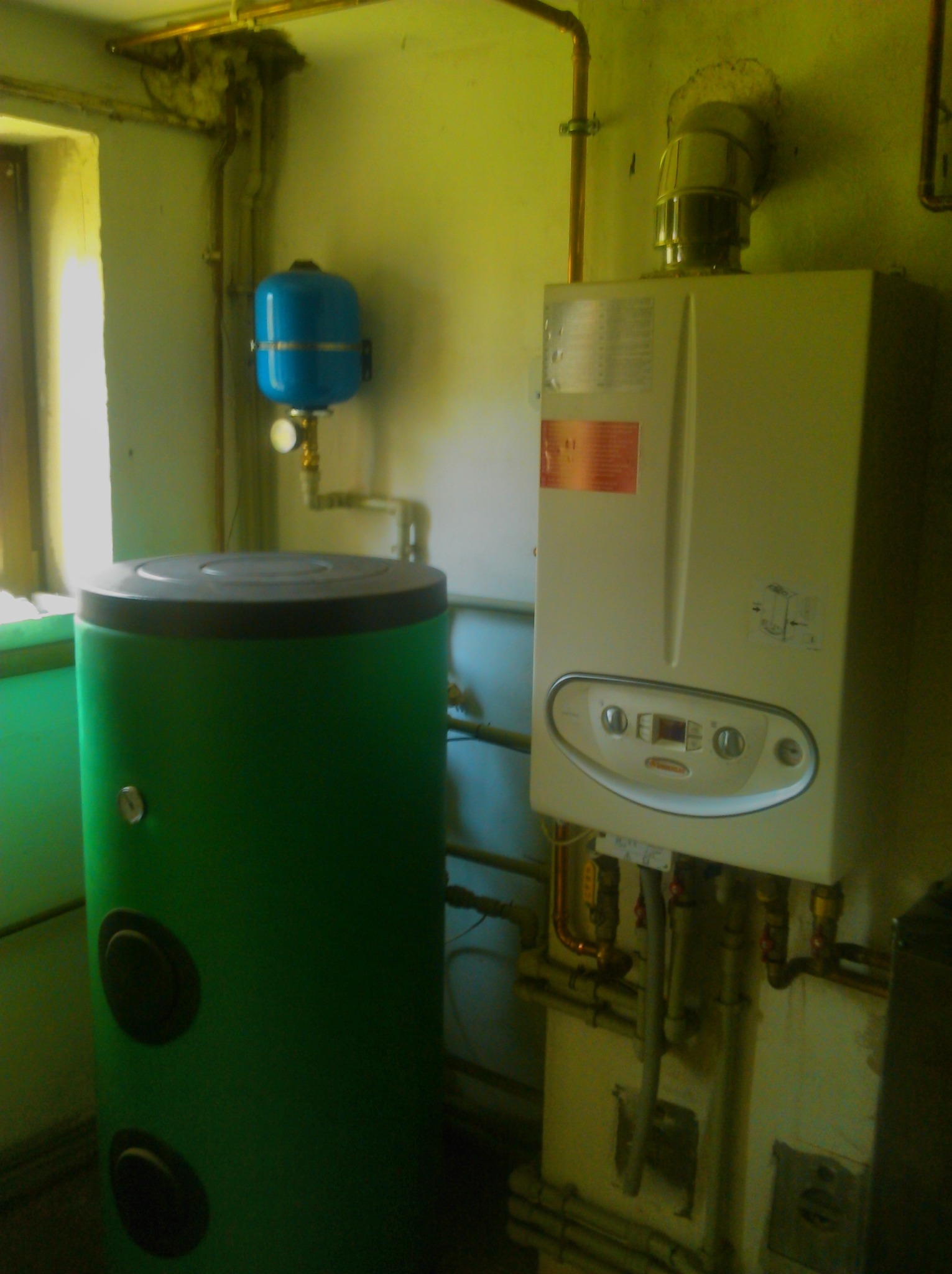 Kocioł gazowy wiszący na ścianie obok pionowego, zielonego zasobnika ciepłej wody i niebieskiego naczynia przeponowego, z widocznymi rurami miedzianymi i instalacją hydrauliczną w pomieszczeniu...