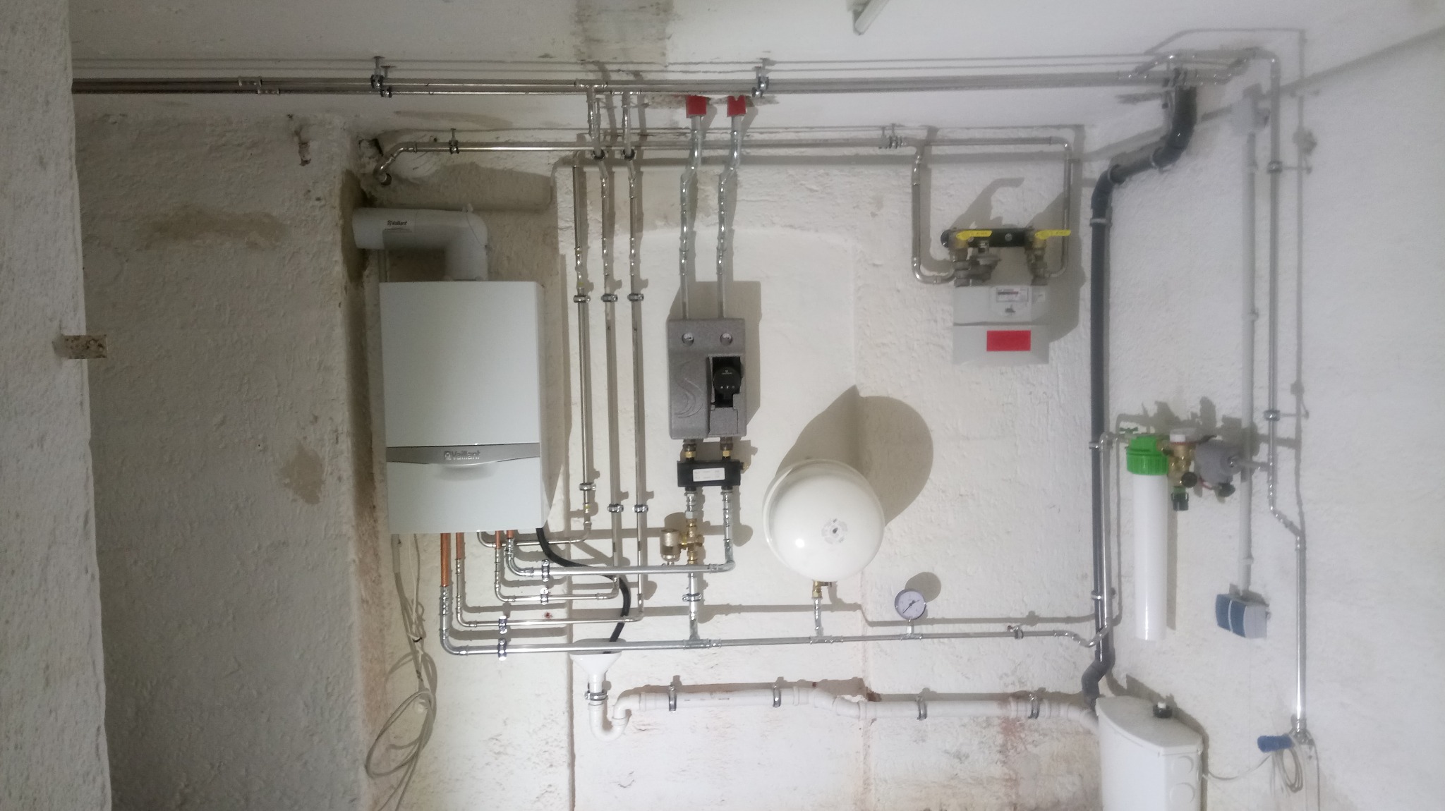 Biały piec gazowy Vaillant zamontowany na szorstkiej ścianie piwnicy z widocznymi rurami miedzianymi i instalacją, w tle licznik gazu i inne elementy instalacji.