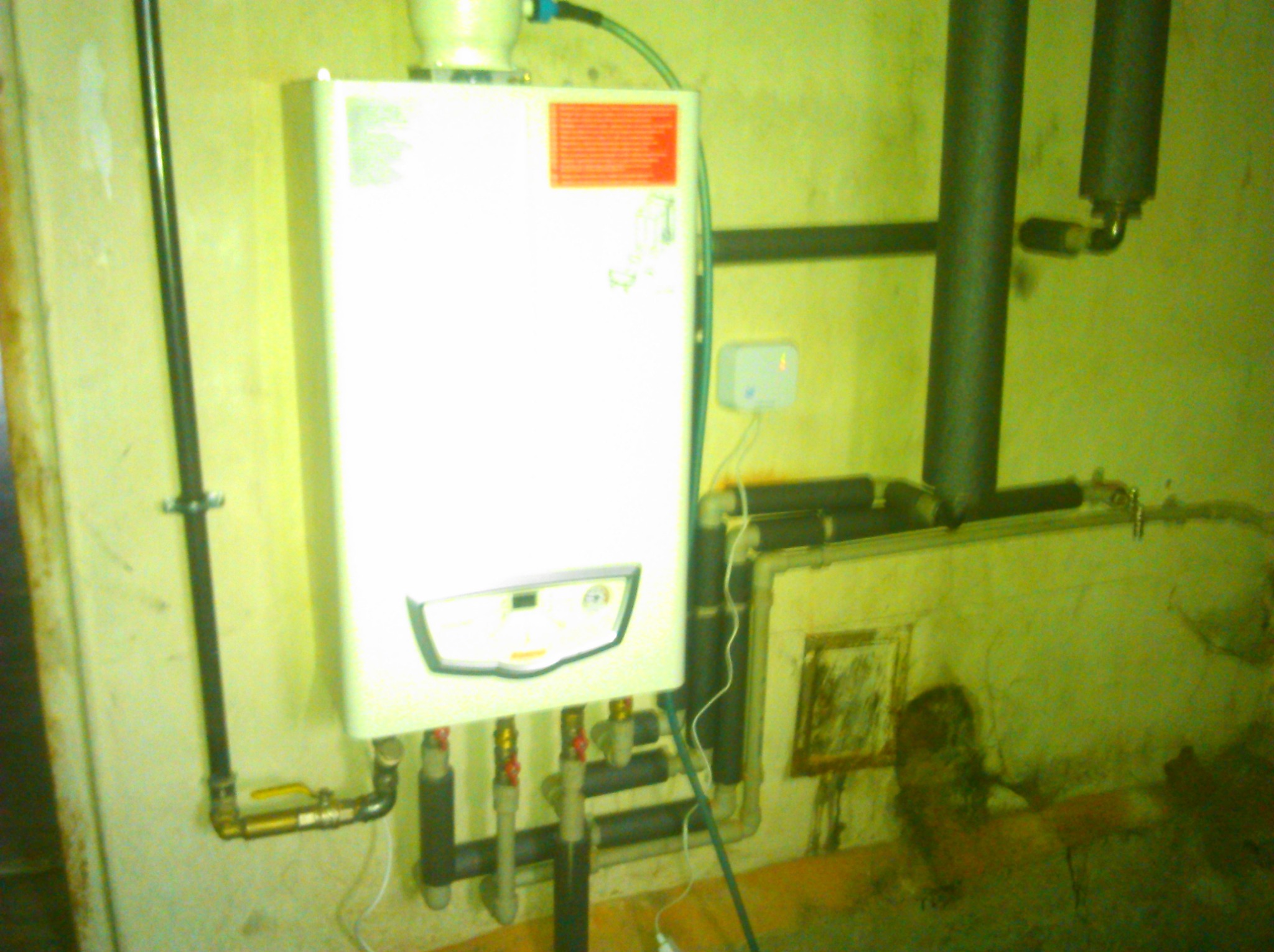 Biały piec gazowy zamontowany na żółtej ścianie, widoczne rury doprowadzające gaz i wodę, oraz instalacja elektryczna. Na piecu czerwona naklejka z informacjami. Nierówności i zabrudzenia na ścianie.