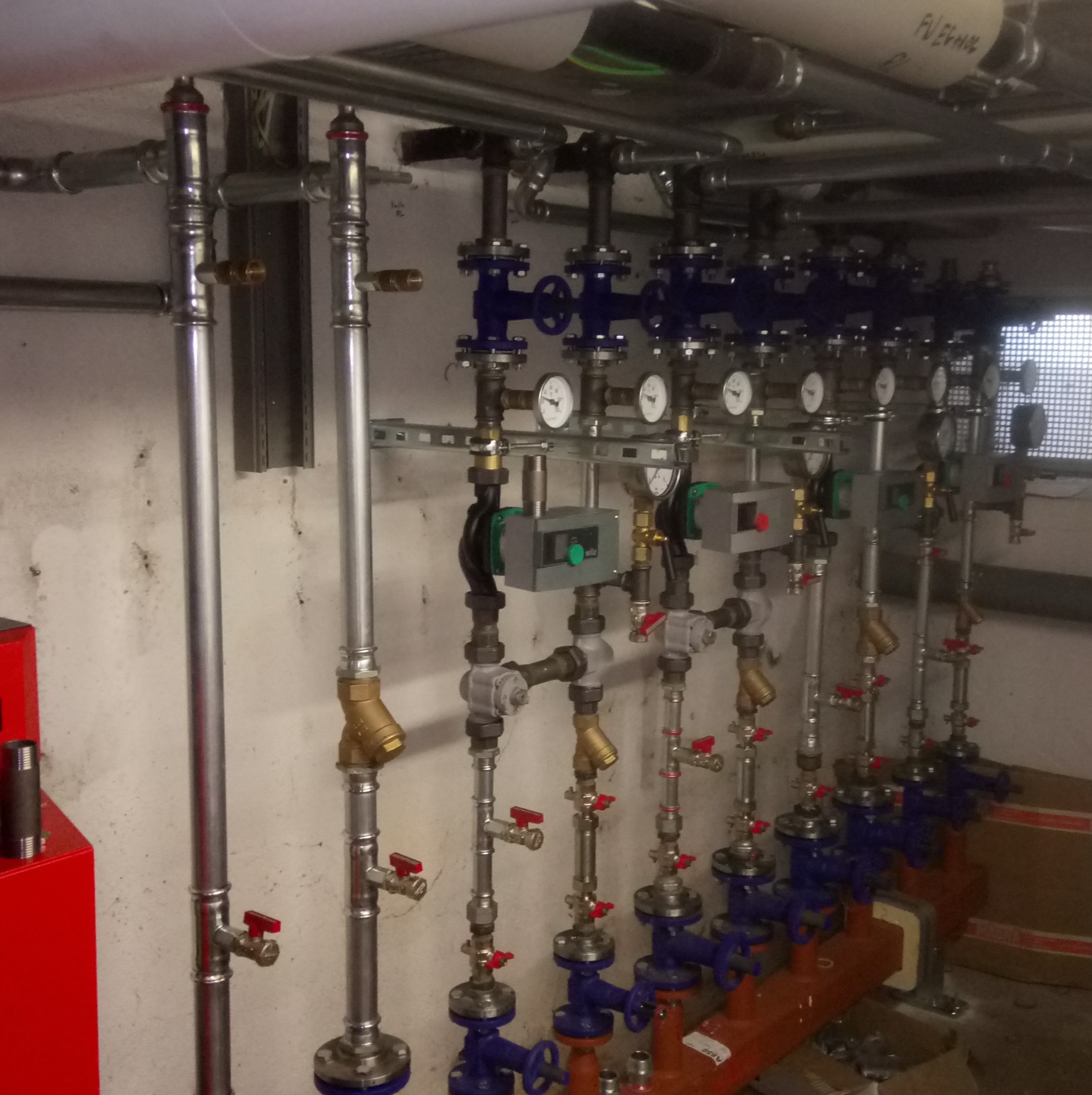 Rzędy pionowych rur instalacyjnych z manometrami i zaworami, niektóre w kolorze niebieskim, zamontowane na ścianie w piwnicy.