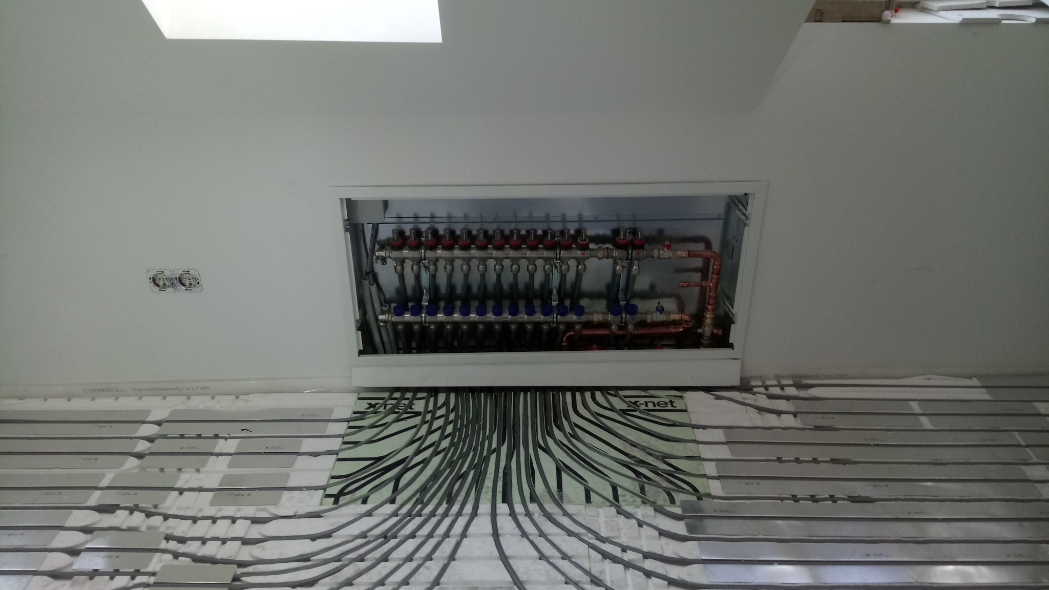 Rozdzielacz ogrzewania podłogowego w zabudowie, widoczne rury rozprowadzone na podłodze z systemem mocowań, instalacja w trakcie montażu.