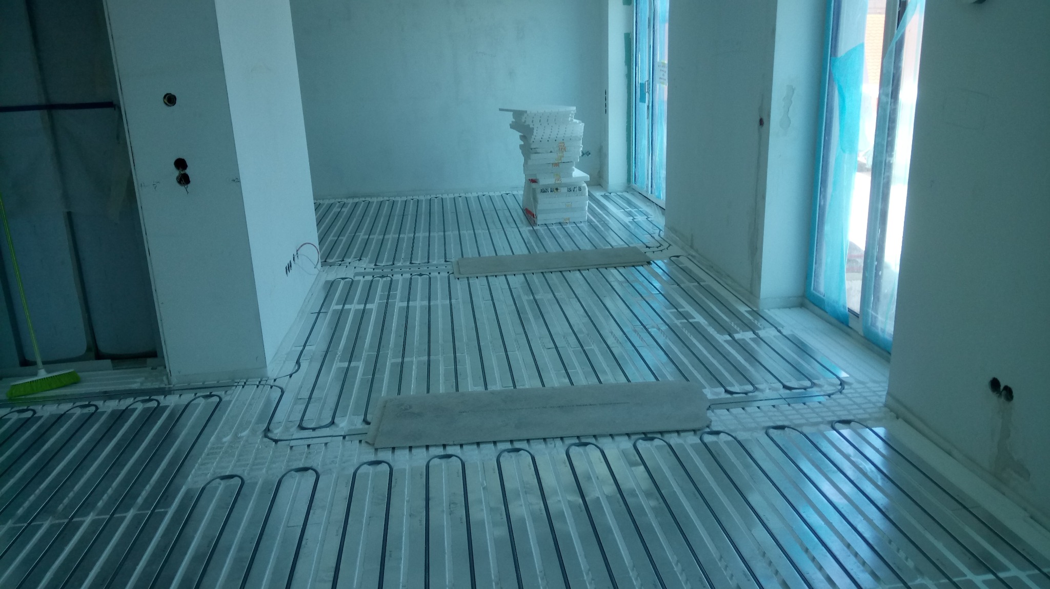 Rozłożone rury ogrzewania podłogowego na izolacji, widoczne płyty izolacyjne, ściany w stanie surowym, okna zabezpieczone folią.