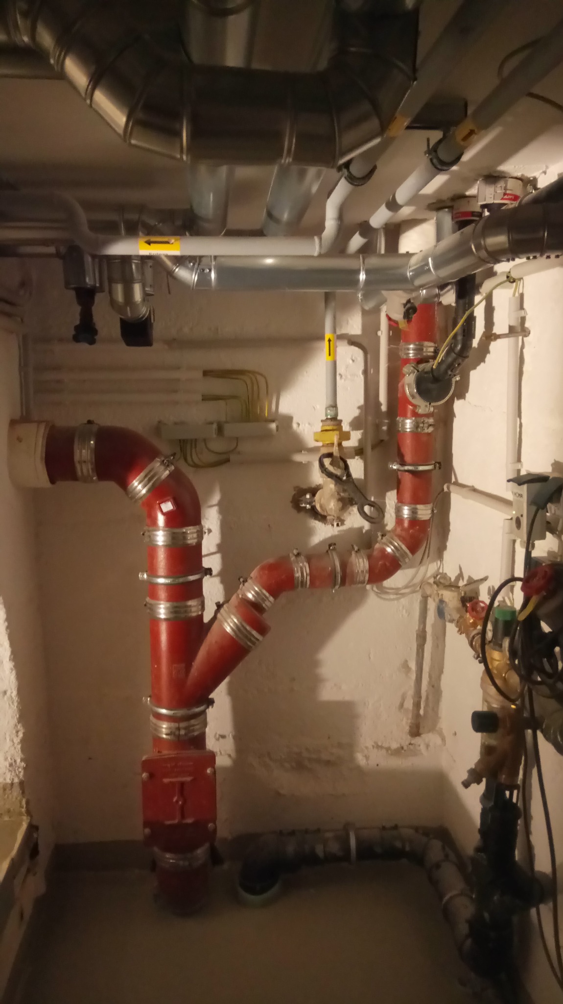 Złożony system rur w piwnicy z czerwonymi rurami połączonymi srebrnymi obejmami, widoczne rury stalowe pod sufitem, żółte strzałki na rurach, instalacja na tle ściany z widoczną strukturą.