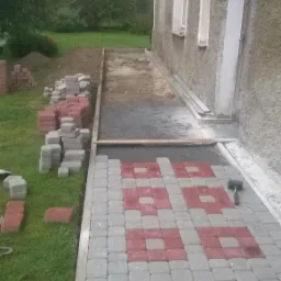 Układanie kostki brukowej w szaro-czerwony wzór na ścieżce przy domu, widoczne przygotowane podłoże i stosy kostki na trawie.