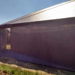 Nowoczesny dom z ciemną elewacją i dachem z dachówki bitumicznej, duże okno narożne, teren wokół domu w trakcie prac ziemnych.