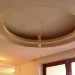 Okrągła dekoracja sufitowa z płyty karton gips, z prowizorycznym oświetleniem w postaci wiszącej żarówki, widok z dołu na sufit w trakcie prac wykończeniowych.