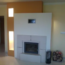 Nowoczesny kominek z obudową z jasnego kamienia, z wkładem kominkowym o ciemnej ramie, narzędzia kominkowe i kosz z drewnem obok, ściana z wnękami oświetleniowymi.