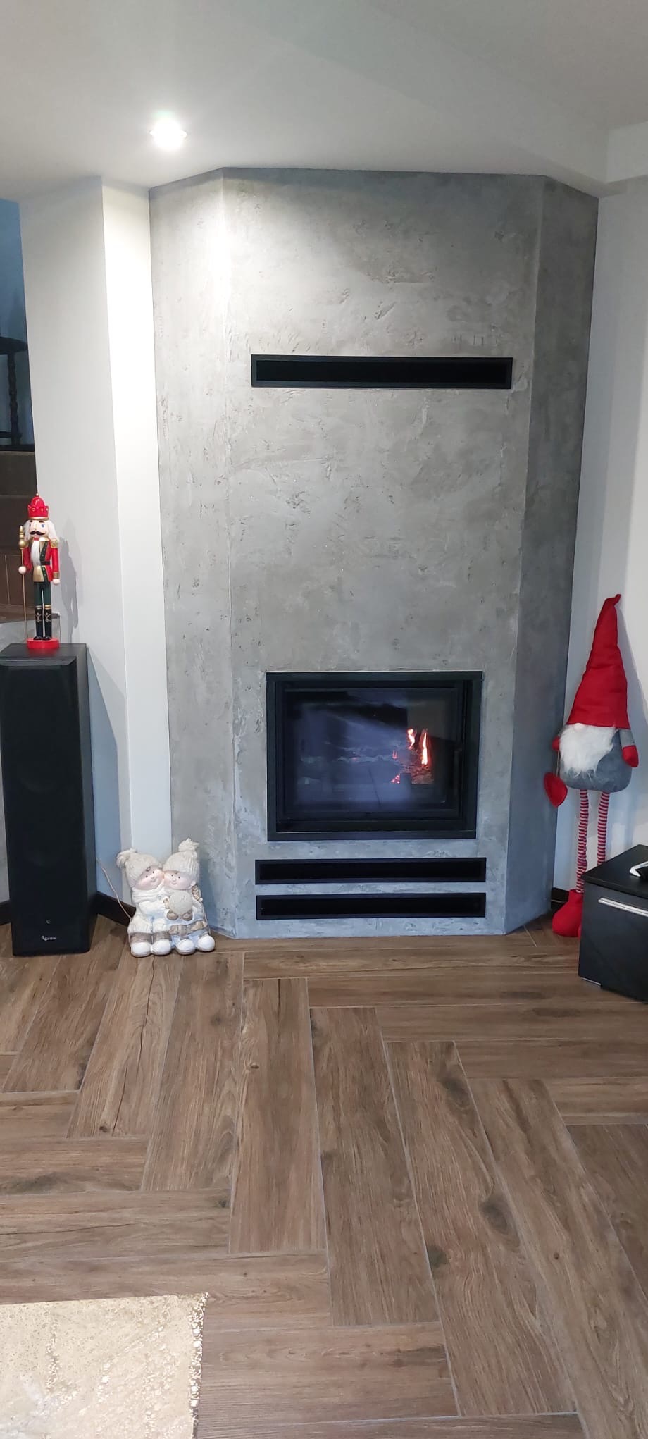 Nowoczesny kominek z szarą obudową o fakturze betonu, z czarnym wkładem i dekoracjami świątecznymi. Parkiet ułożony w jodełkę. Wnętrze domu.