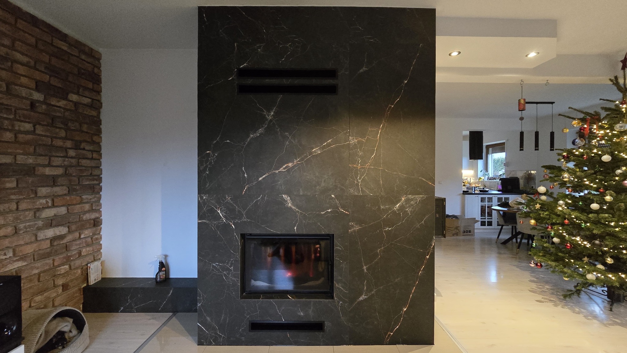 Nowoczesny kominek z czarnego marmuru z widocznym paleniskiem, w tle choinka i wnętrze domu. Dekoracja świąteczna w salonie z ceglaną ścianą.