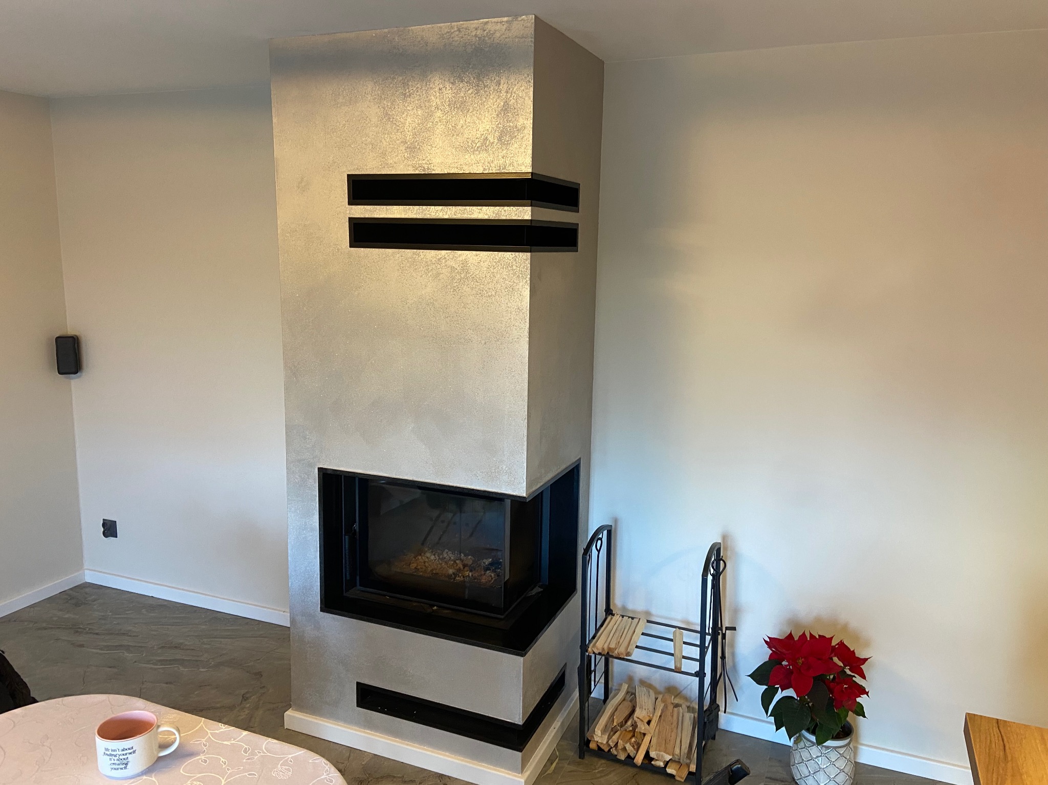 Nowoczesny kominek narożny z obudową w odcieniach szarości, z czarnymi akcentami, drewnem i poinsecją, tworzący przytulną atmosferę w jasnym wnętrzu.