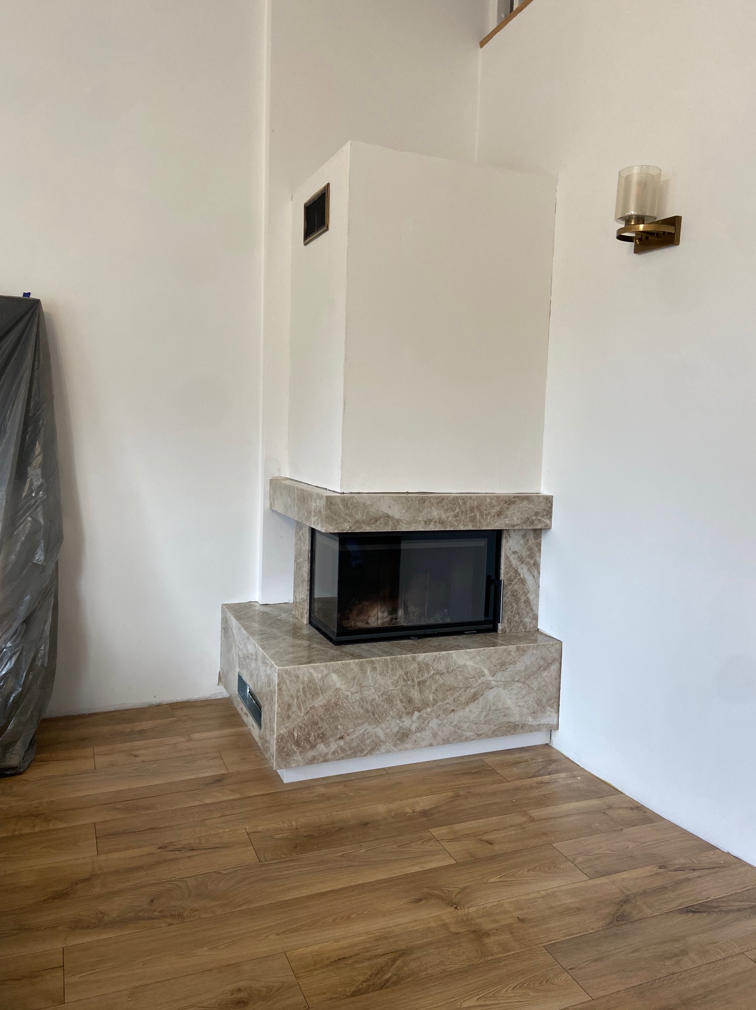 Nowoczesny kominek z marmurową obudową i czarnym wkładem, na tle białej ściany z kinkietem. Widoczna drewniana podłoga i zabezpieczona folią część pomieszczenia.