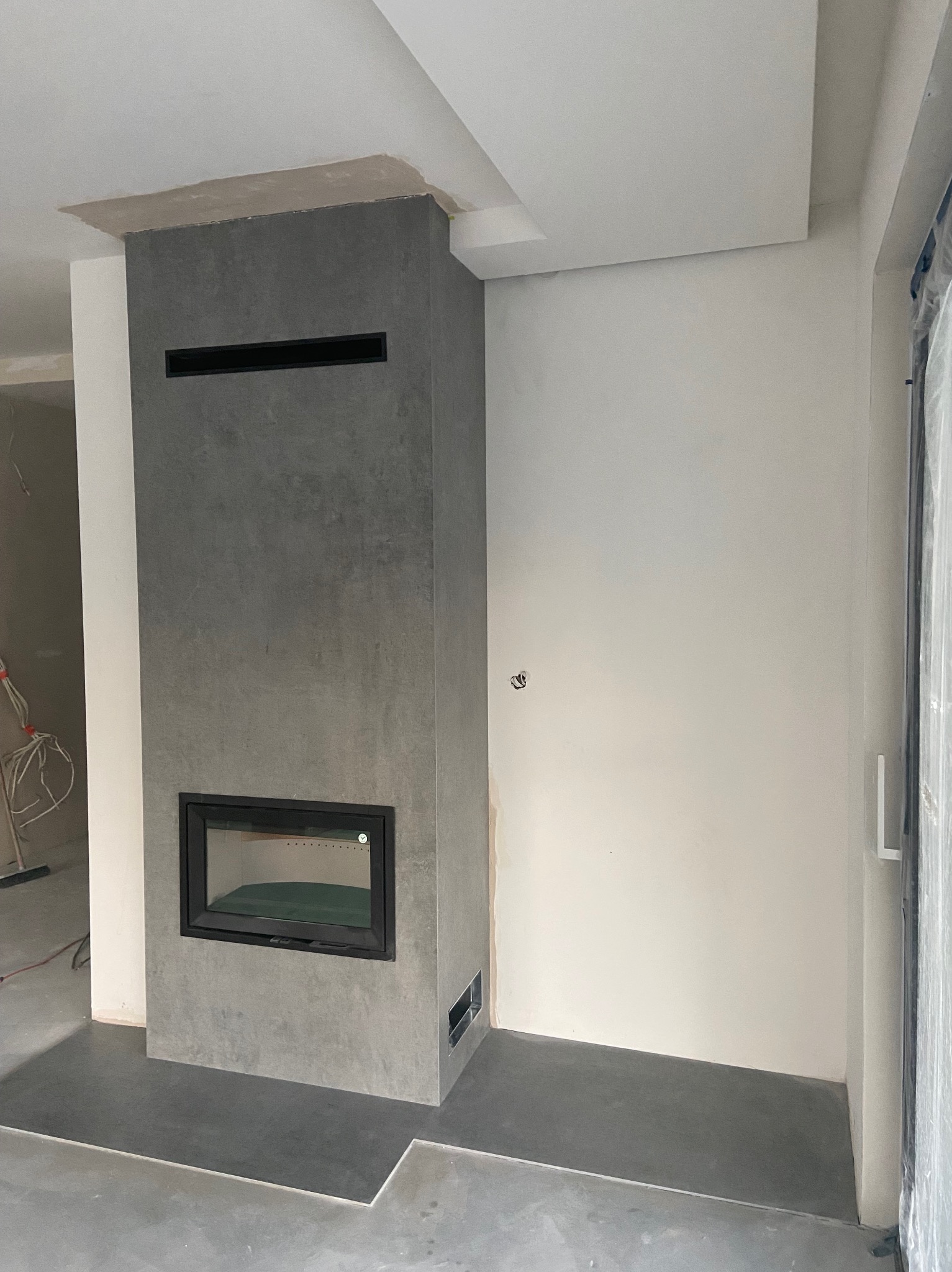 Nowoczesny kominek z szarego kamienia z czarną obudową, w trakcie montażu. Widoczne niedokończone prace wykończeniowe na suficie i podłodze.