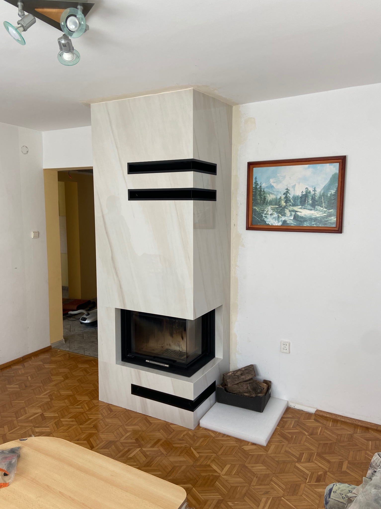 Nowoczesny kominek z obudową z jasnego marmuru, z czarnymi akcentami, w jasnym wnętrzu z parkietem i obrazem górskim na ścianie. Widoczne drewno do palenia.