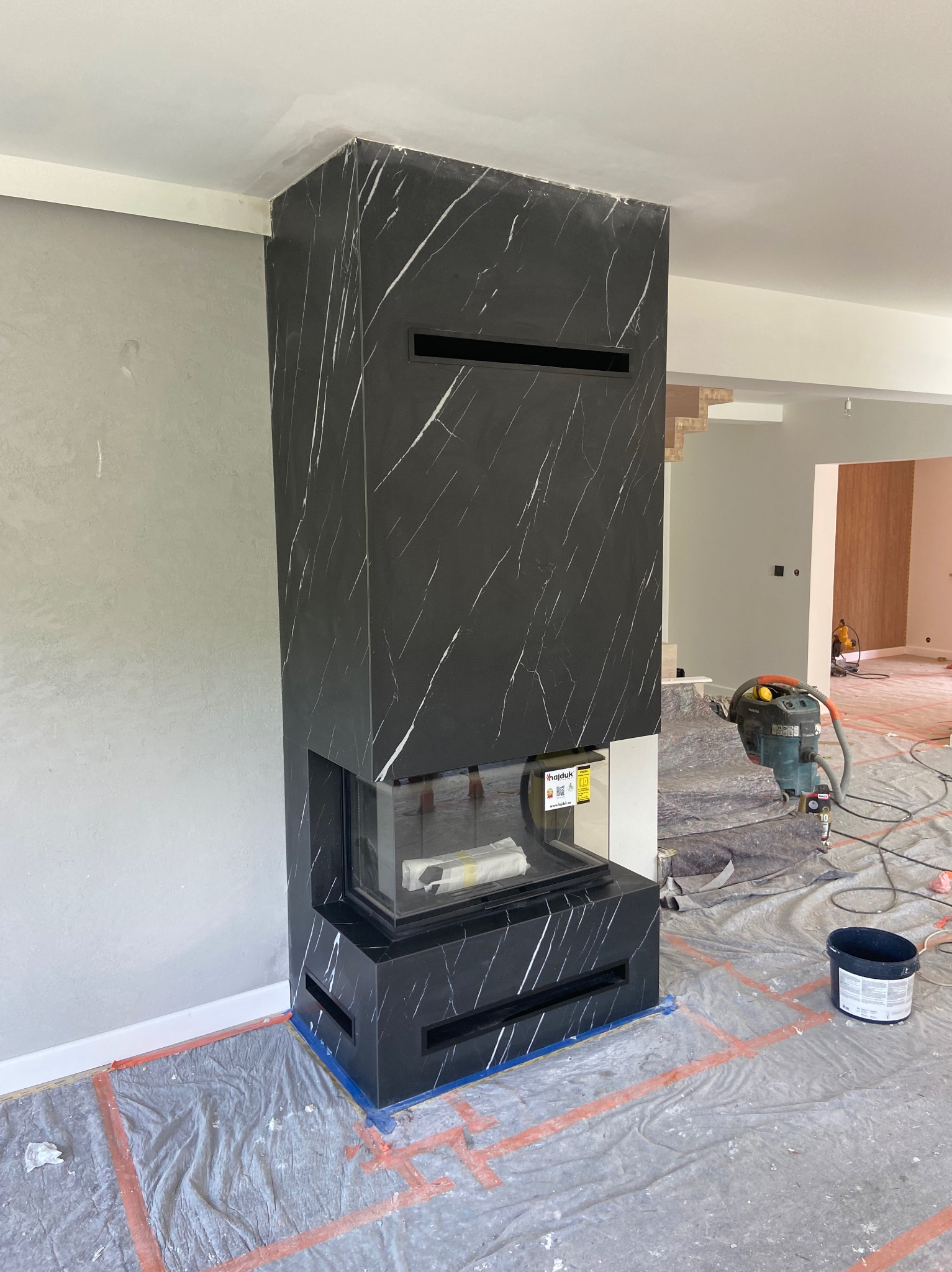 Nowoczesny kominek z czarnego marmuru z białymi żyłami, w trakcie montażu. Widoczne narzędzia i zabezpieczenia podłogi. Minimalistyczny design, surowe wykończenie ścian.
