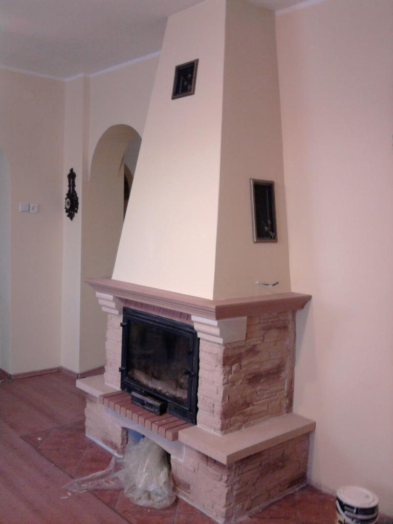 Wykończony kominek z obudową z cegły i czarnymi drzwiczkami, na podłodze widoczny worek z odpadami i farba, ściany w kolorze kremowym.