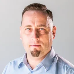 Portret mężczyzny w błękitnej koszuli z kozia bródką na jasnoszarym tle.