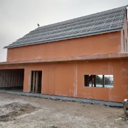 Surowy stan domu szkieletowego z pomarańczową elewacją, widoczne elementy konstrukcyjne dachu i otwory okienne, teren wokół w trakcie przygotowania.