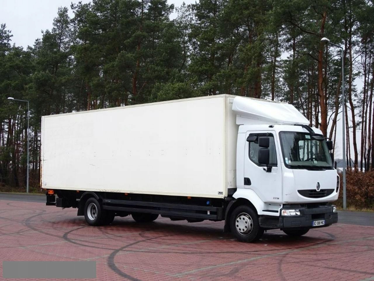 Biały samochód dostawczy Renault z zabudową kontenerową na parkingu z czerwonej kostki, widok z boku na tle lasu.