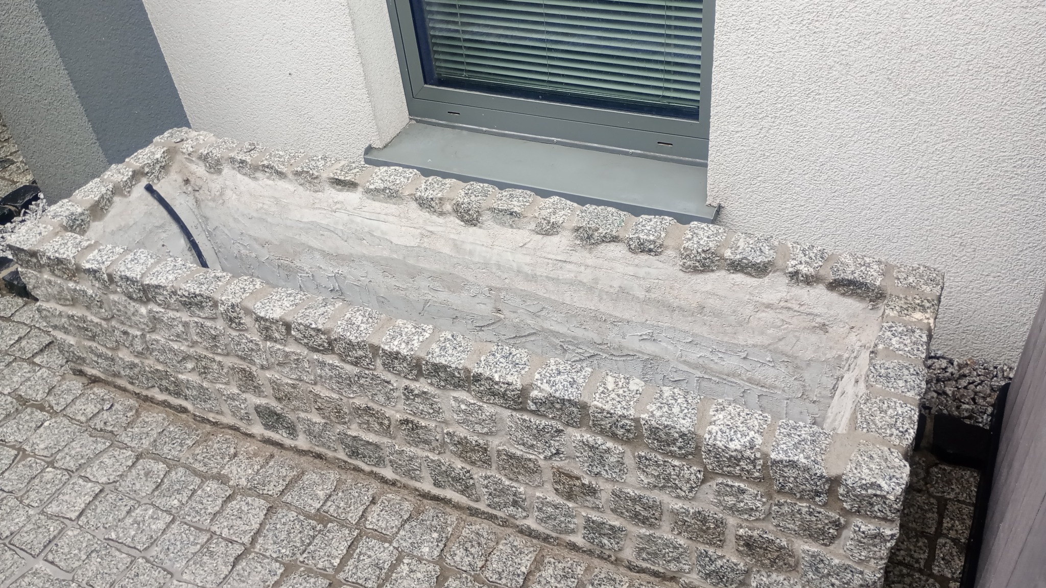 Granitowa donica pod oknem, wyłożona kostką. Widoczna tekstura kamienia i szara tonacja. Donica w surowym stanie, bez roślin. Kostka granitowa na podłożu.