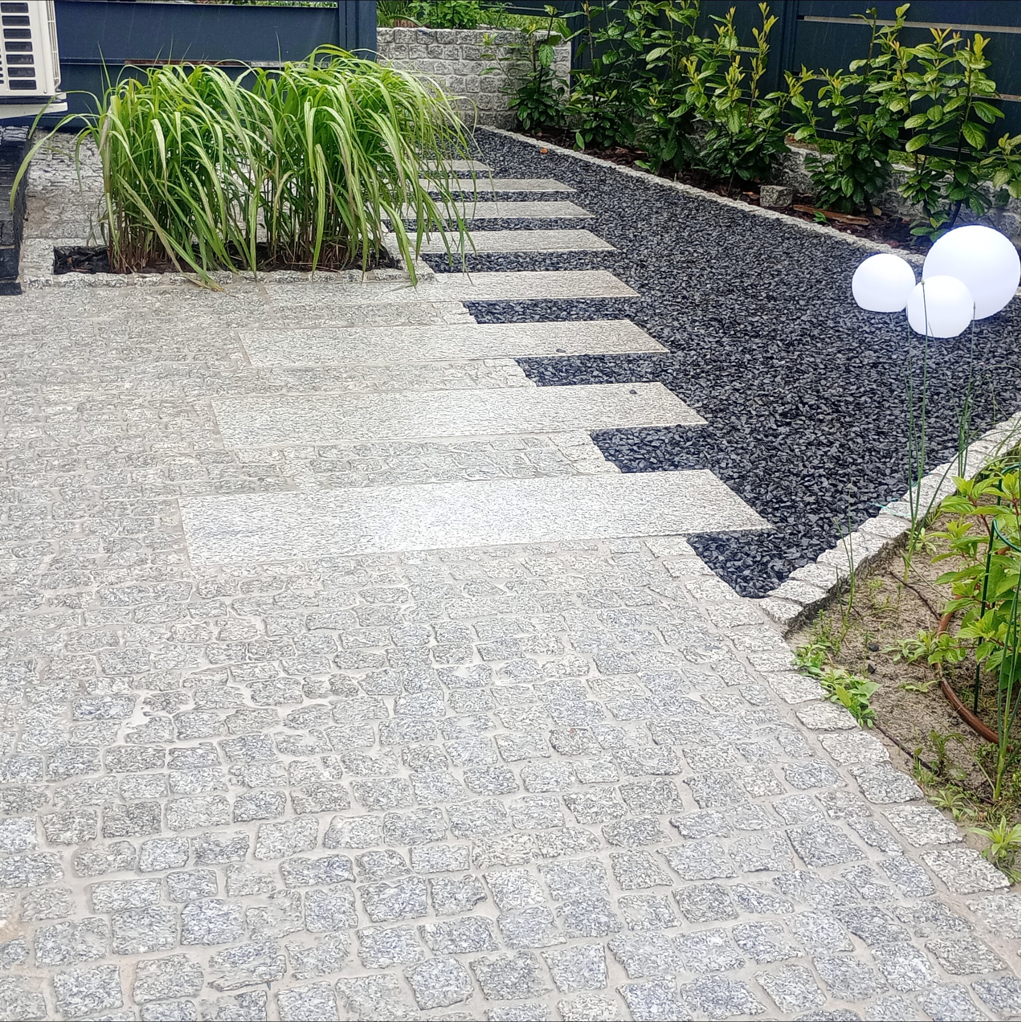 Granitowa ścieżka z elementami dekoracyjnymi: roślinność i lampy kuliste. Układanka z kostki granitowej przechodzi w płytki i czarny żwir.