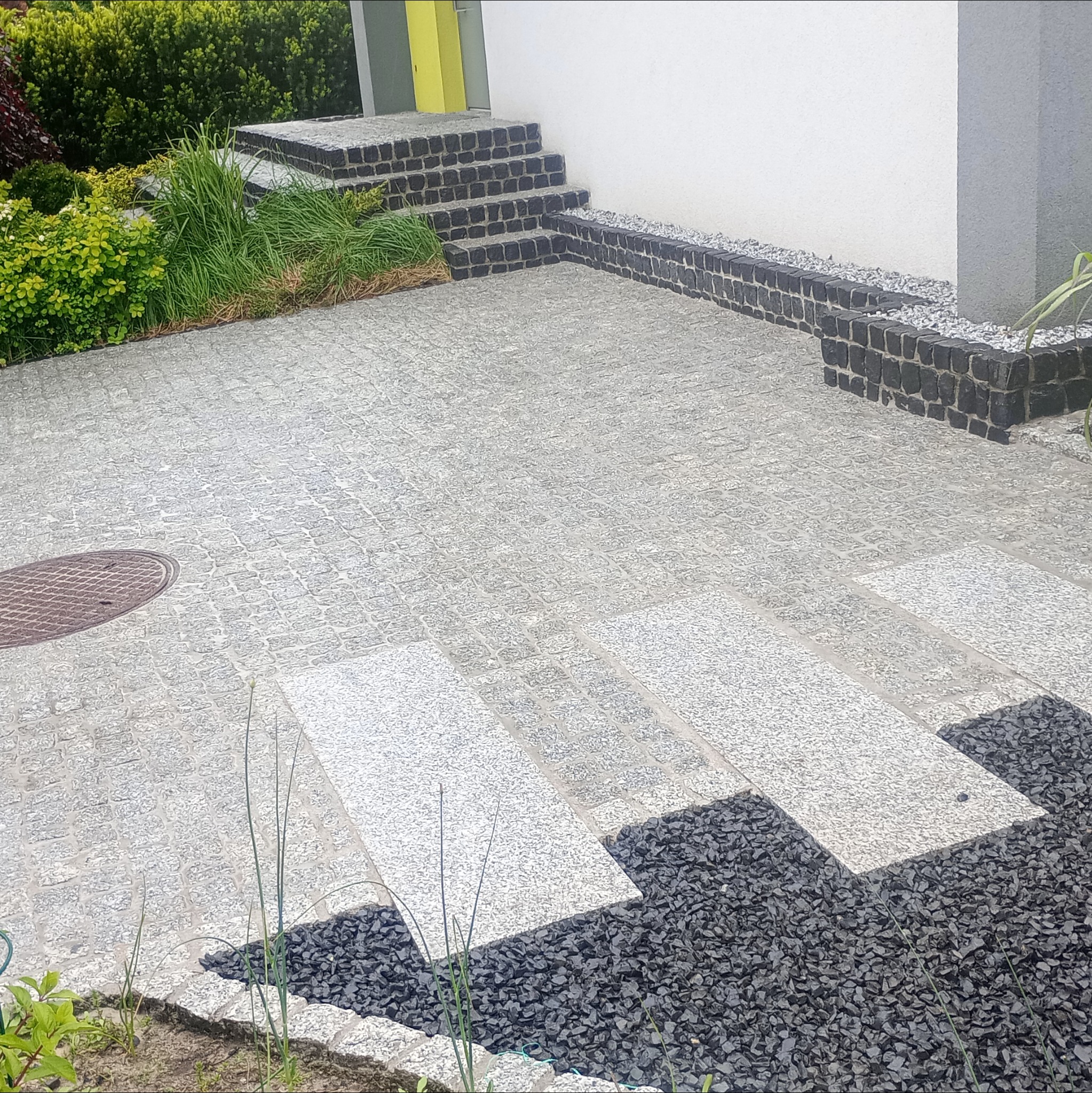 Ułożona jasnoszara kostka granitowa tworzy ścieżkę z ciemnymi akcentami z kamienia, prowadzącą do schodów z kostki i żółtych drzwi wejściowych. Widoczne rośliny.