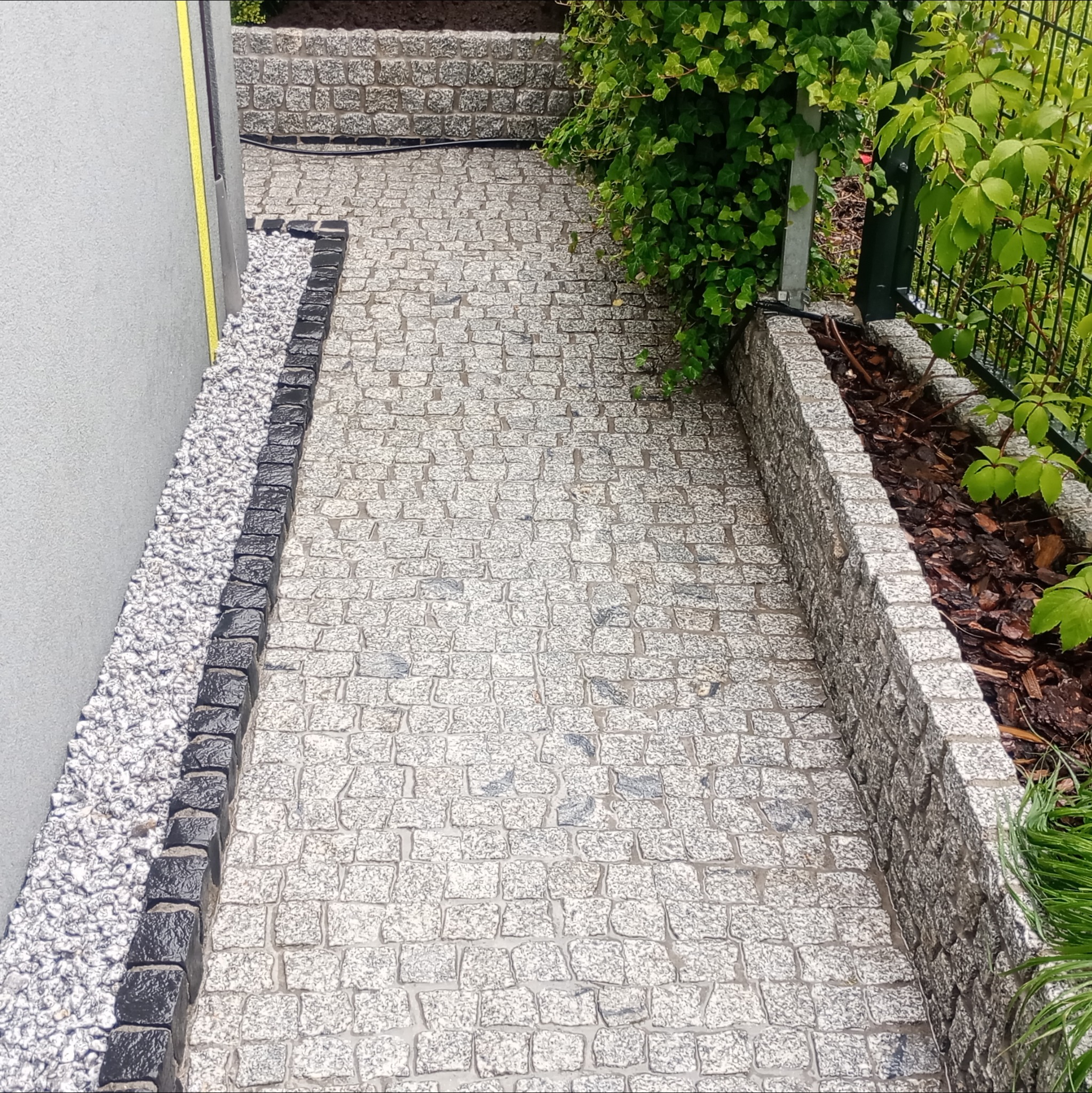 Ścieżka z kostki granitowej z czarnym obramowaniem, obok ściany z białym żwirem i rabatą z roślinami. Widok z góry.