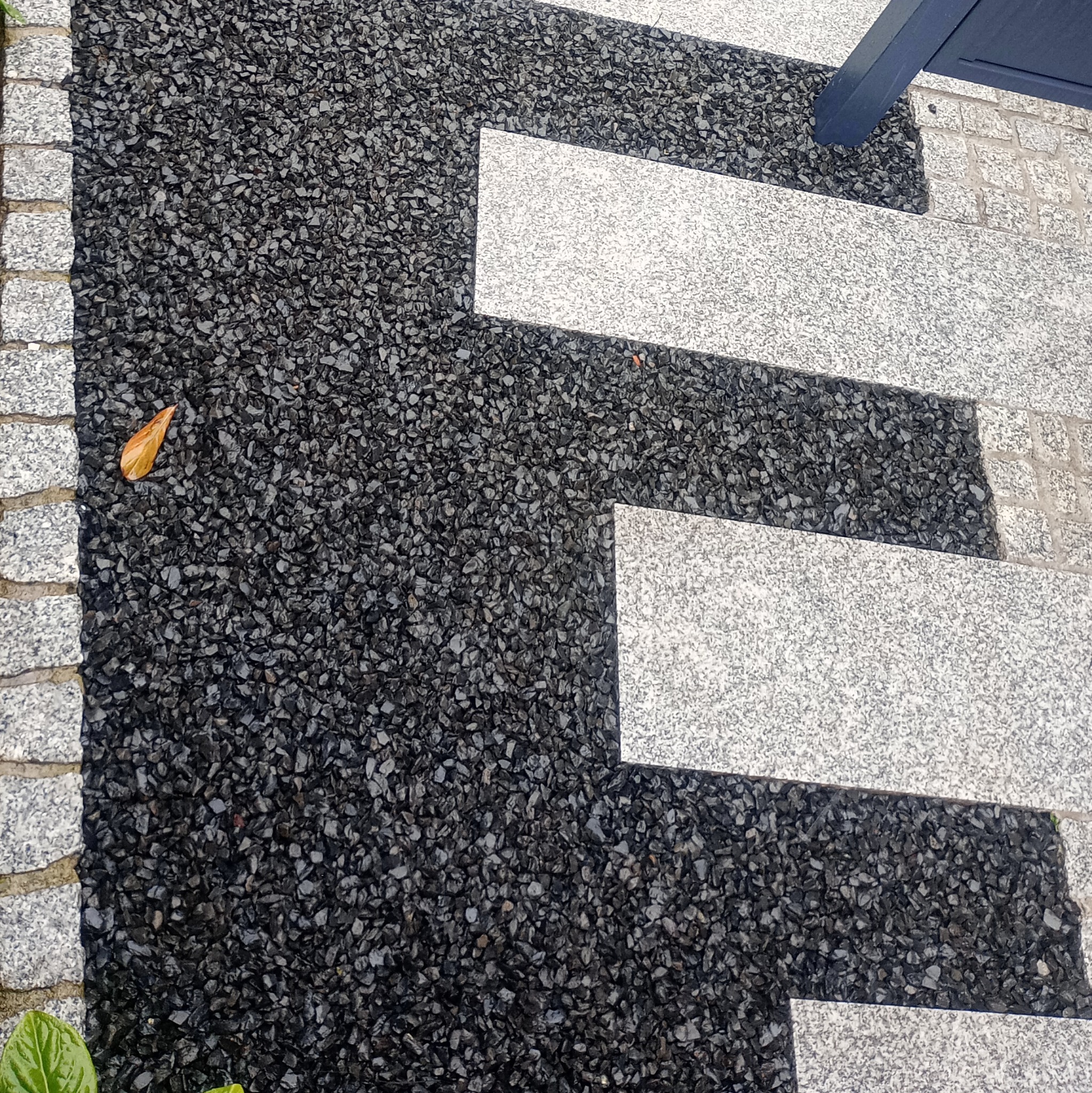 Ścieżka w ogrodzie z ciemnego żwiru i jasnych, prostokątnych płyt kamiennych. Brzeg wykończony kostką. Widoczny opadły liść i fragment rośliny.