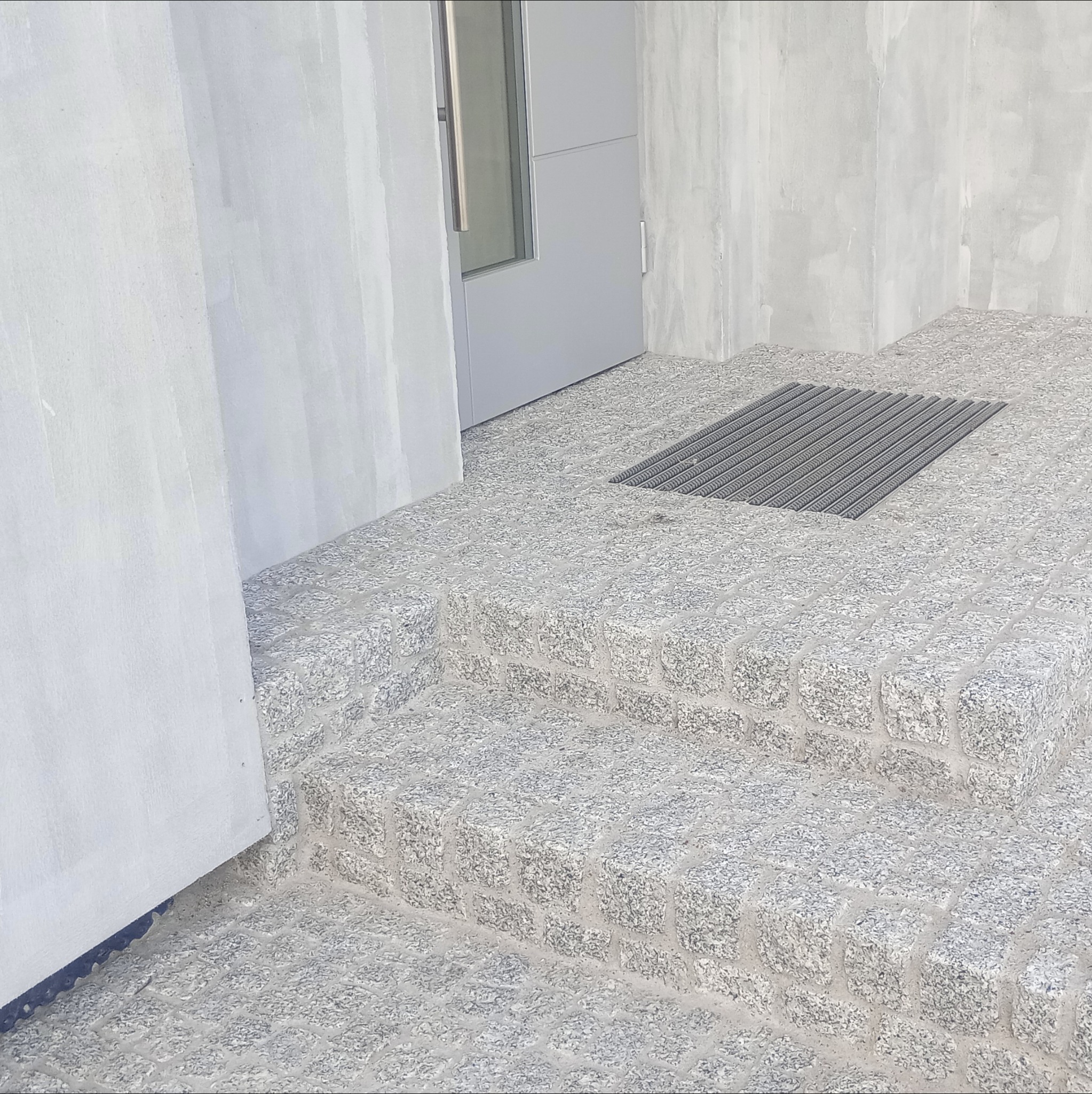 Granitowe schody wejściowe z kratką odpływową, prowadzące do nowoczesnych drzwi. Jasnoszara kostka tworzy solidną i estetyczną powierzchnię.