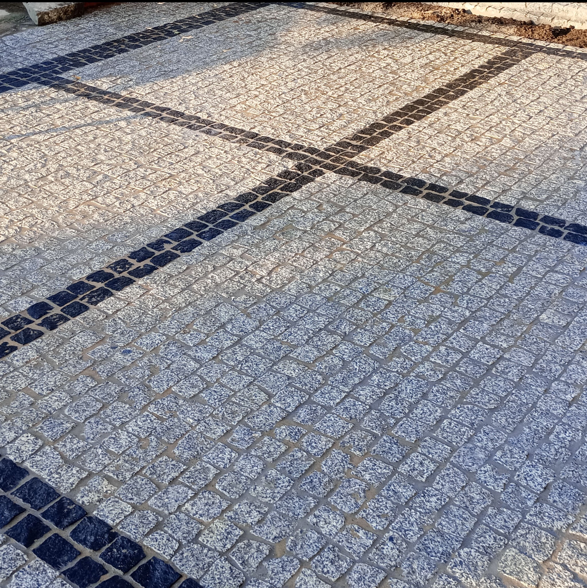 Ułożona kostka granitowa w odcieniach szarości i błękitu, z ciemnymi pasami tworzącymi wzór krzyża. Widok z góry, ukazujący regularność i precyzję wykonania.