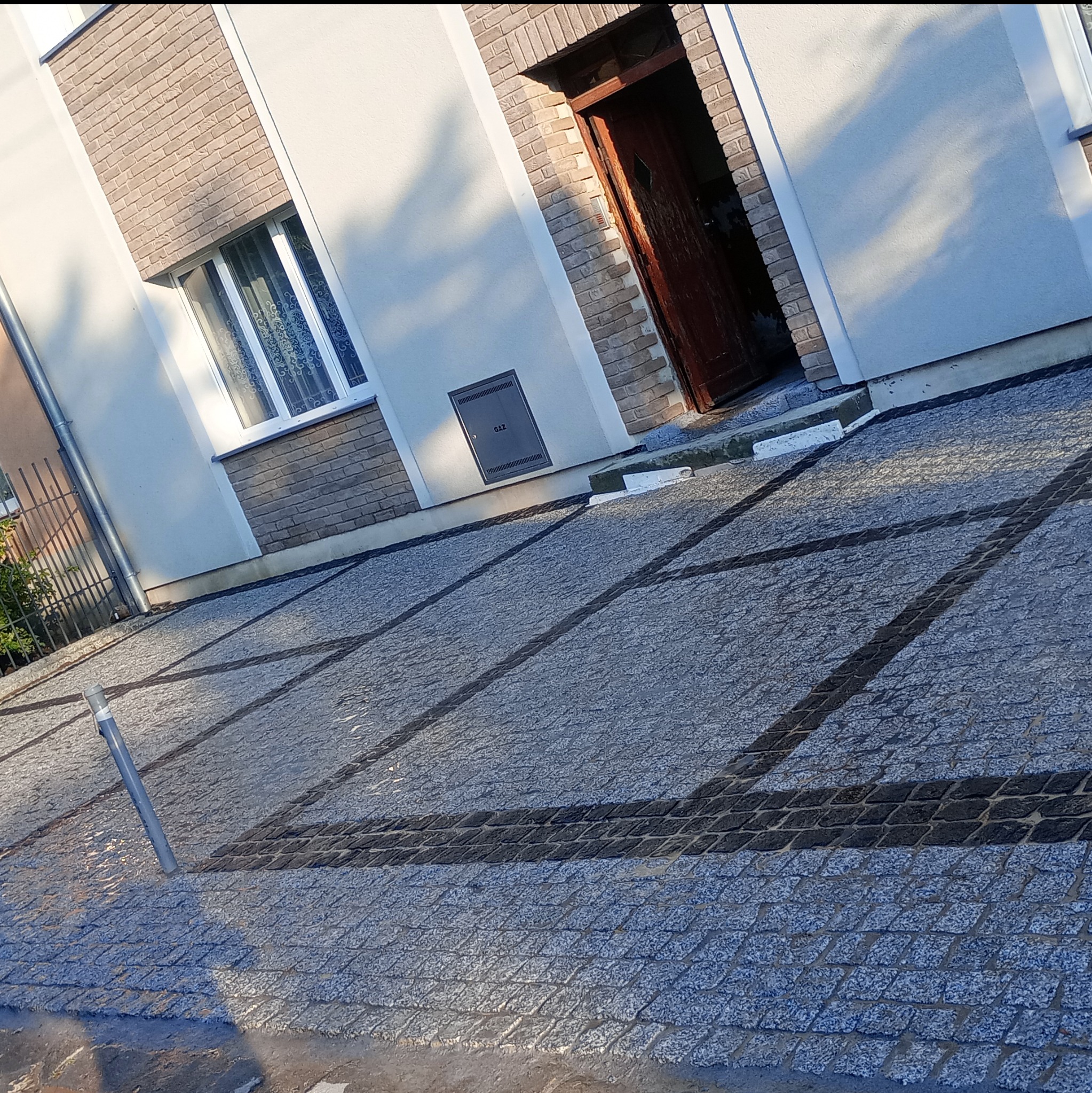 Ułożona kostka granitowa na podjeździe przed domem z oknem i drewnianymi drzwiami wejściowymi. Widoczne ciemne pasy kostki oddzielające obszary.
