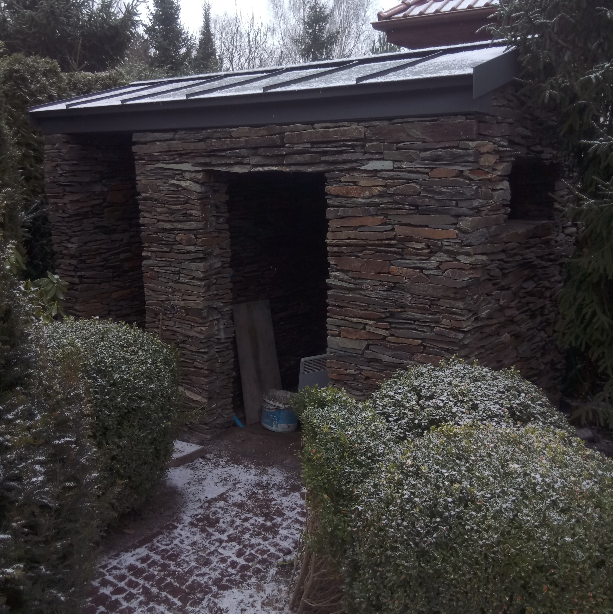 Kamienna altana z zadaszeniem, otoczona zimozielonymi krzewami, częściowo pokryta śniegiem. Widoczne narzędzia i materiały budowlane wewnątrz.