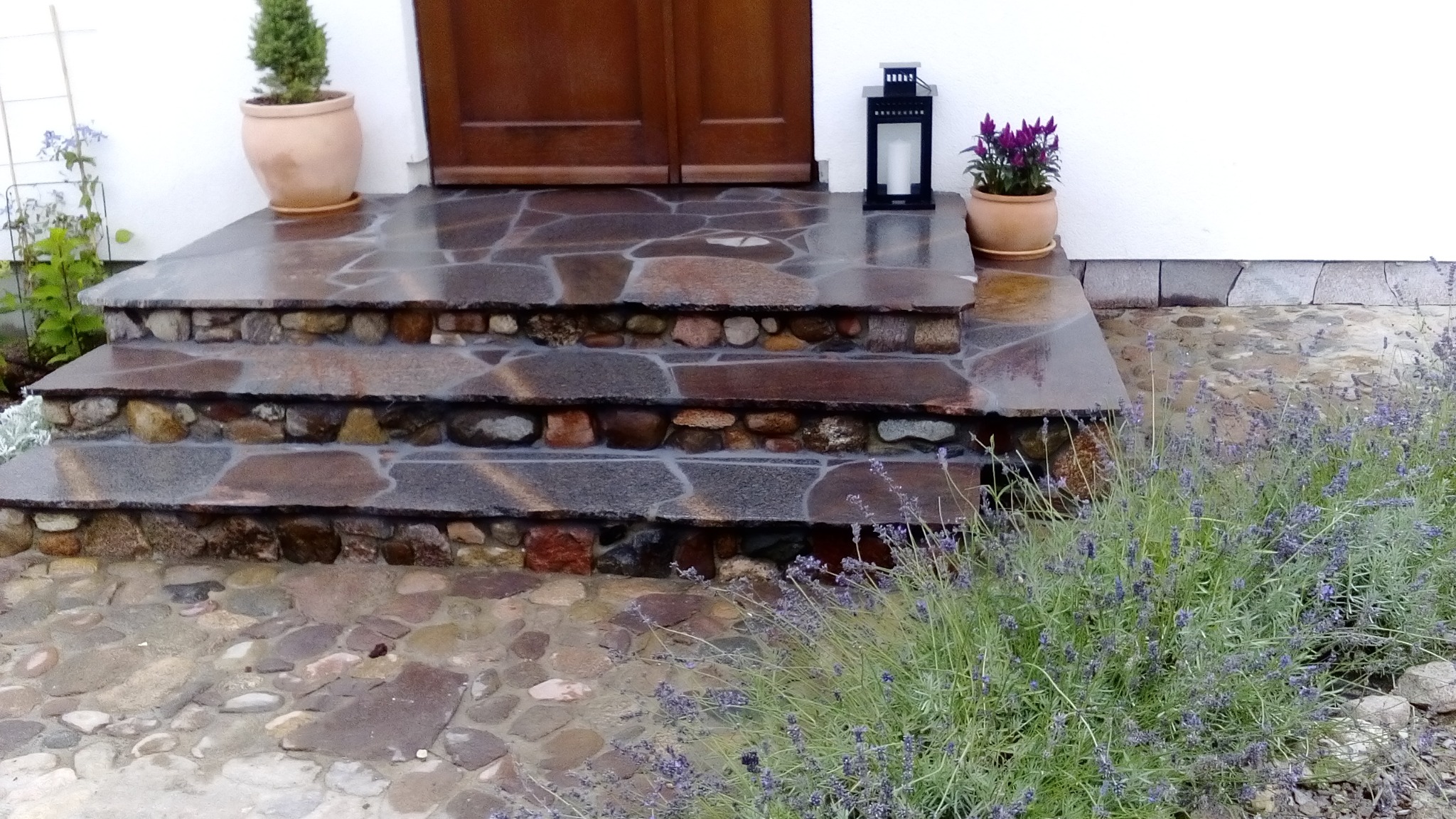 Kamienne schody wejściowe z roślinami w donicach i ozdobnym lampionem, wykończone kamieniem naturalnym, prowadzą do drewnianych drzwi wejściowych.