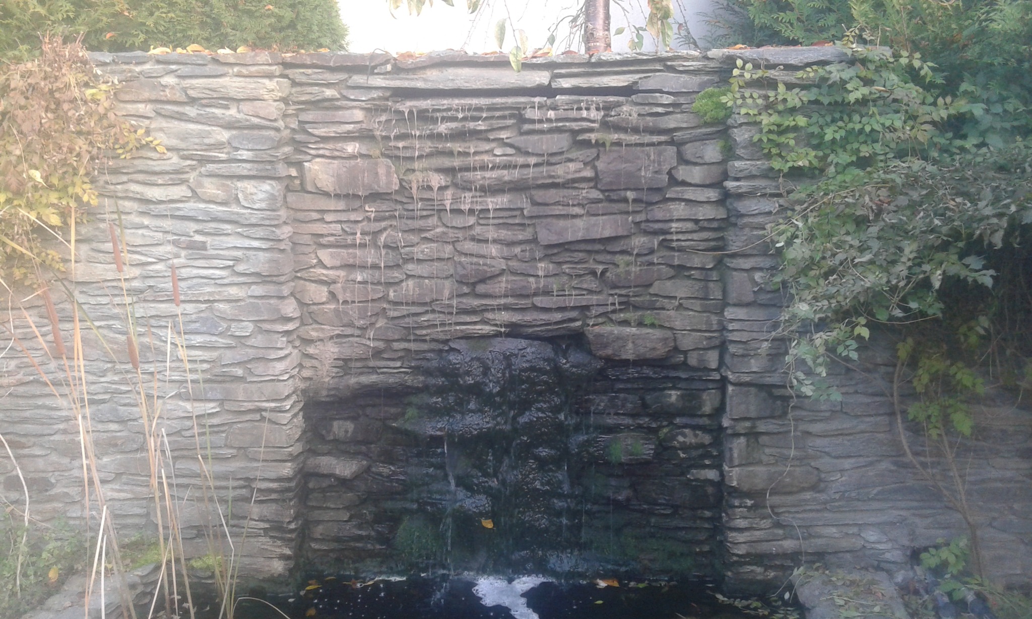 Kamienna kaskada wodna w ogrodzie, wykonana z płaskich kamieni, otoczona zielenią i trzciną. Woda spływa po kamieniach do małego stawu u podstawy.