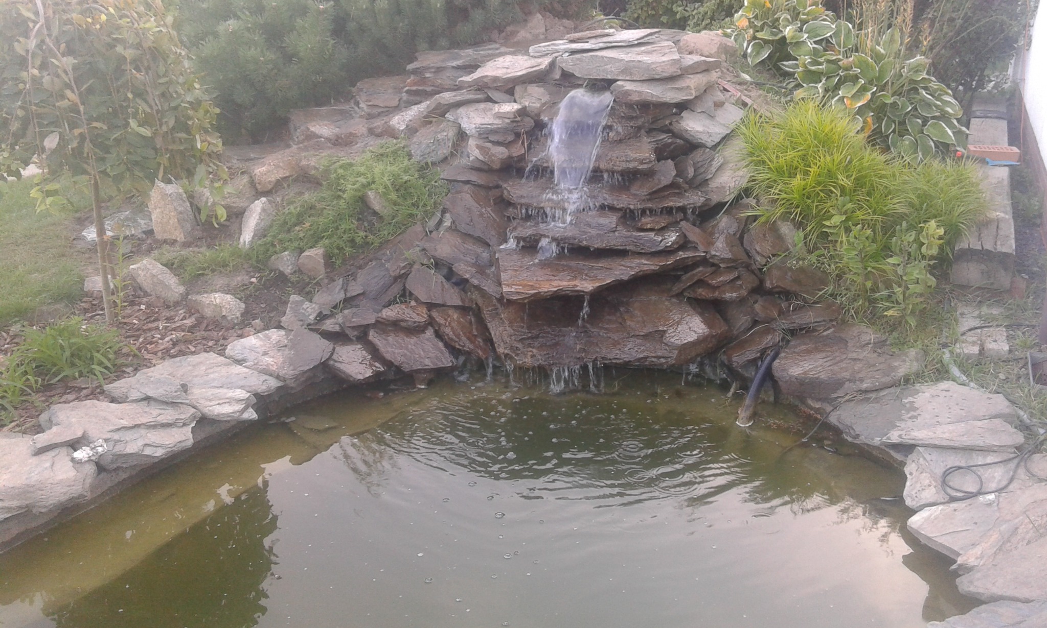 Kaskada wodna z kamienia łupkowego, zasilana rurą, spływająca do oczka wodnego otoczonego zielenią i kamieniami. Ujęcie z bliska, w naturalnym oświetleniu.