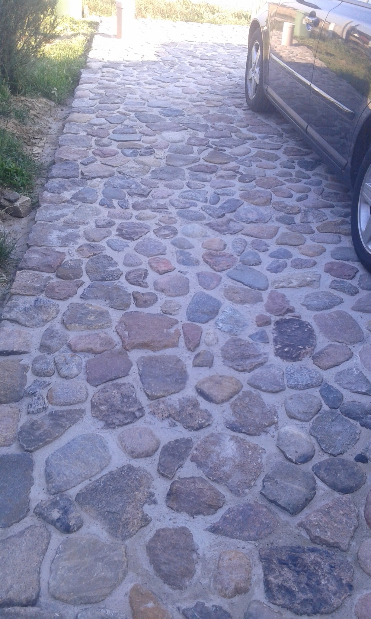 Kamienna ścieżka z nieregularnych kamieni, obok zaparkowany samochód. Ścieżka prowadzi w głąb kadru, otoczona zielenią i fragmentami trawy. Widok z góry, oświetlenie naturalne.
