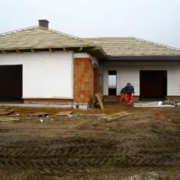 Budowa domu jednorodzinnego w trakcie ocieplania ścian styropianem, widoczne rusztowania dachowe i niezagospodarowany teren wokół, dwóch pracowników w roboczych ubraniach.