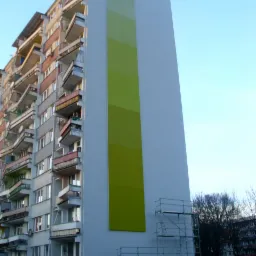 Wysoki blok mieszkalny z fragmentem elewacji w odcieniach zieleni, widoczne rusztowania i zaparkowane samochody u podnóża budynku.