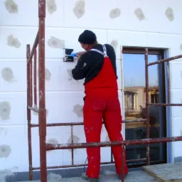 Pracownik w czerwonym kombinezonie, stojąc na rusztowaniu, montuje puszkę elektryczną w ocieplonej styropianem ścianie budynku, widoczne zaślepione otwory po kołkach.