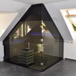 Szklana sauna wkomponowana w skos dachu, z drewnianymi ławkami i podświetleniem LED. Uwagę zwraca nowoczesny design i dopasowanie do architektury wnętrza.