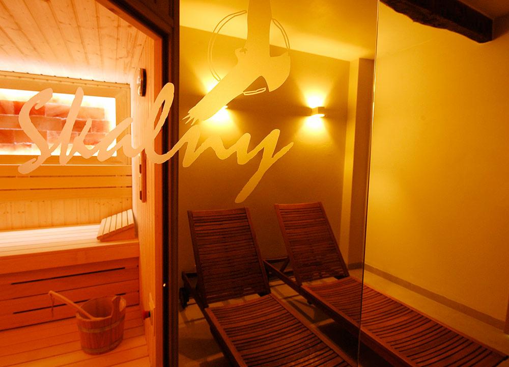 Wnętrze sauny z drewnianymi leżakami, widoczne przez szklane drzwi z napisem. Oświetlenie ciepłym światłem.