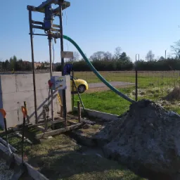 Proces wiercenia studni: wiertnica z niebieskim silnikiem, zielony wąż odprowadzający urobek, sterta ziemi obok wykopu, w tle ogrodzenie i żółty samochód.