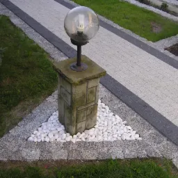 Oświetlenie ogrodowe z kulistą lampą na kamiennym postumencie, otoczone białymi otoczakami, na tle ścieżki z jasnej kostki z ciemnymi obrzeżami.