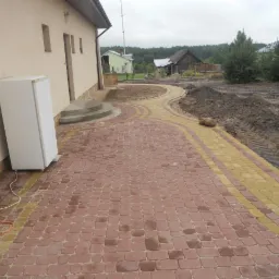 Nowo ułożony chodnik z czerwono-żółtej kostki brukowej prowadzący do domu, z widocznymi śladami opon na ziemi obok i lodówką stojącą przy ścianie budynku.