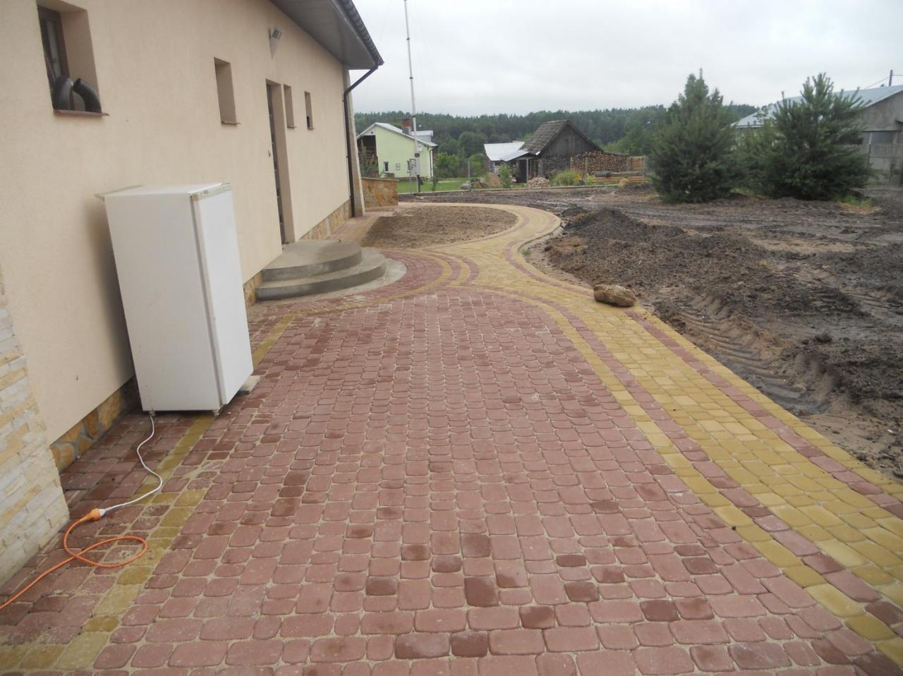 Nowo ułożony chodnik z czerwono-żółtej kostki brukowej prowadzący do domu, z widocznymi śladami opon na ziemi obok i lodówką stojącą przy ścianie budynku.